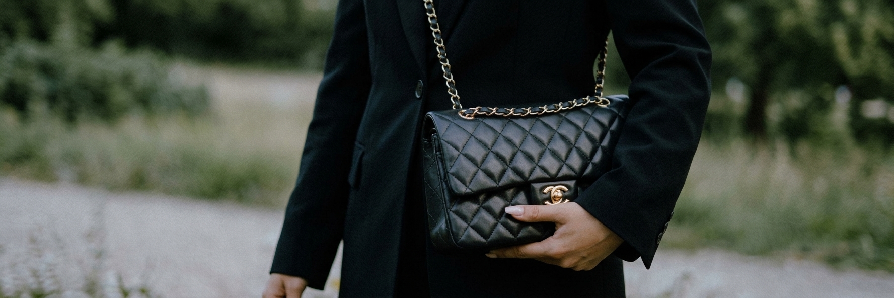 A person in a black coat holding a quilted black handbag with a gold chain strap outdoors.