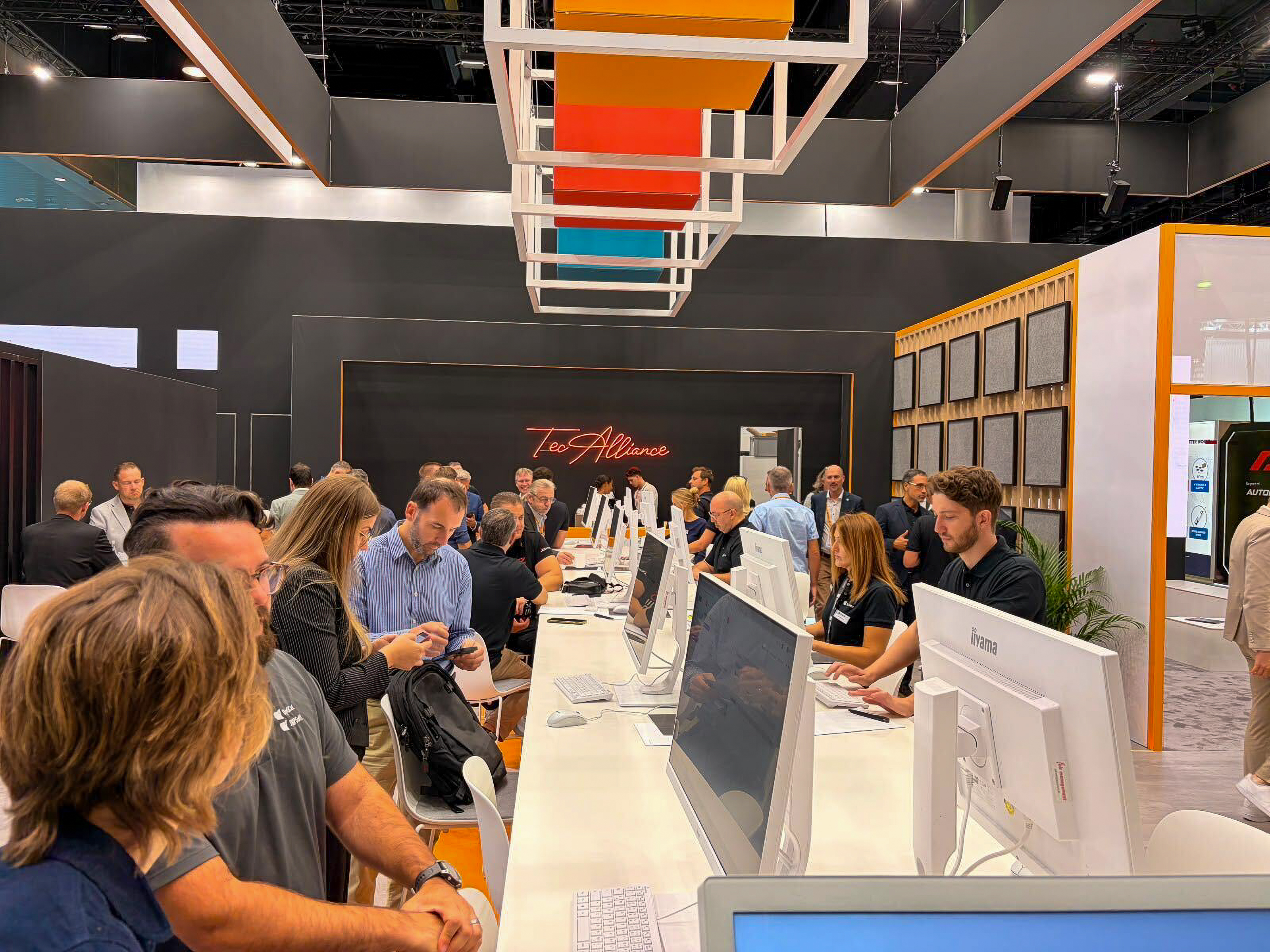 A busy booth at Automechanika Frankfurt with people interacting at a long desk, using computers and discussing, with colorful overhead decor and a black background.