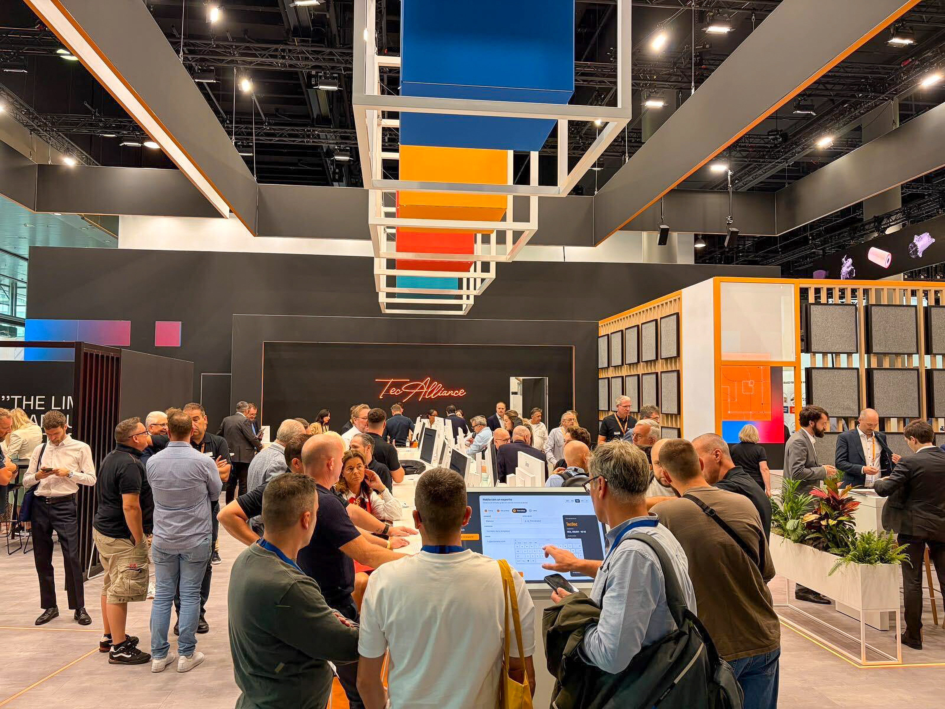 TecAlliance booth at Automechanika Frankfurt 2026 A busy exhibition booth with people interacting and using digital screens, beneath colorful ceiling structures, in a modern trade fair setting.