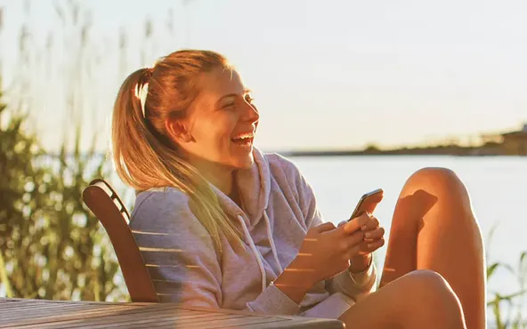 Ein Mann und eine Frau sitzen bei Sonnenuntergang an einem See, lächeln und halten Smartphones in der Hand.