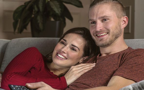 Ein Paar sitzt lächelnd auf einer Couch und sieht fern. Die Frau stützt ihren Kopf auf die Schulter des Mannes, der eine Fernbedienung hält. Im Hintergrund steht eine Pflanze.