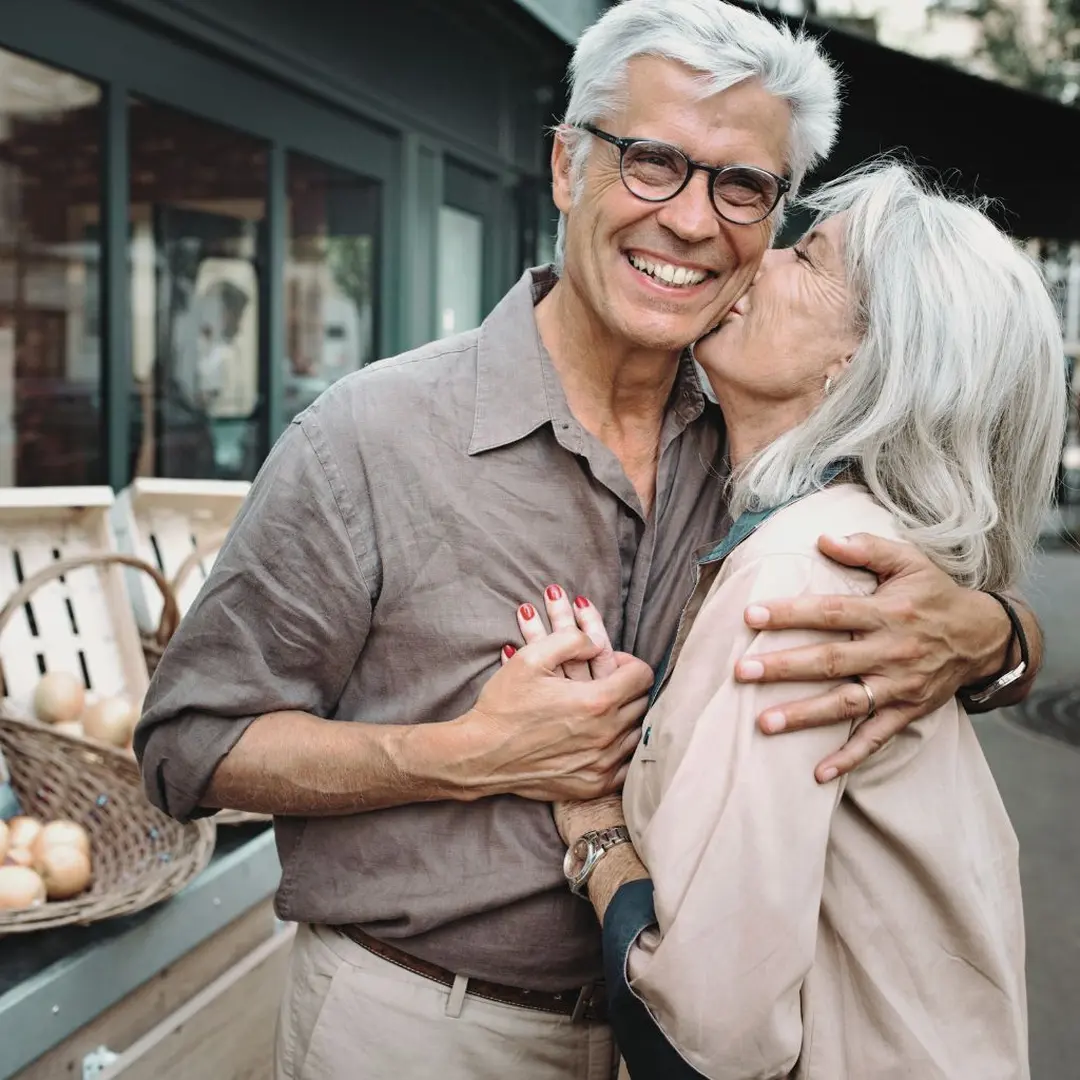 An older couple embraces cheerfully outdoors, with the woman giving the man a kiss on the cheek while he smiles broadly.