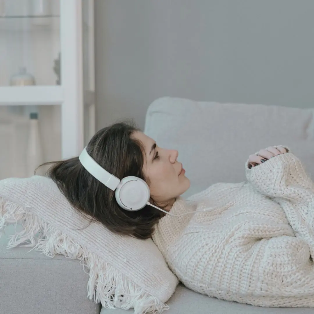 A woman wearing headphones and a knit sweater lies on a sofa, resting her head on a cushioned pillow.