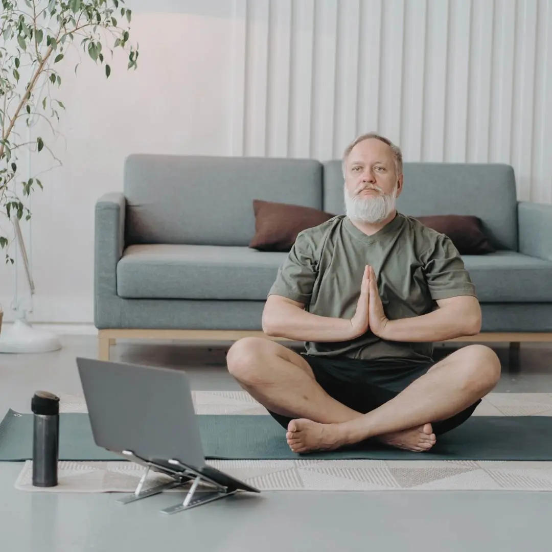 Man practicing yoga in a living room, seated cross-legged on a mat with hands in prayer position, facing a laptop. A couch and plant are in the background.