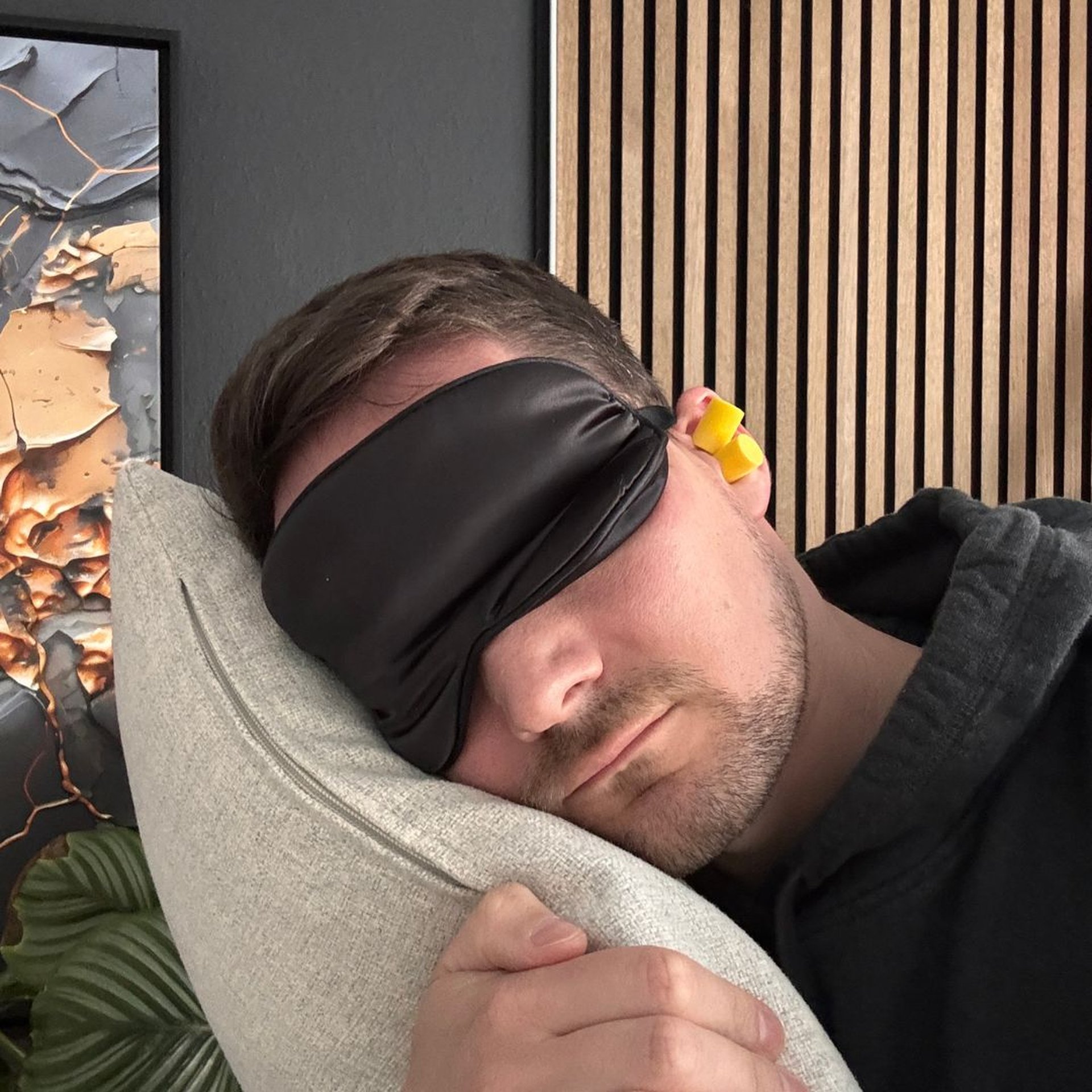 Man sleeping on a pillow, wearing a black sleep mask and yellow earplugs, with a textured wall and plant in the background.