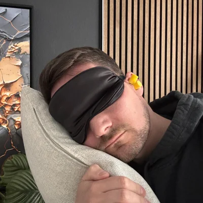 Man sleeping on a pillow, wearing a black sleep mask and yellow earplugs, with a textured wall and plant in the background.
