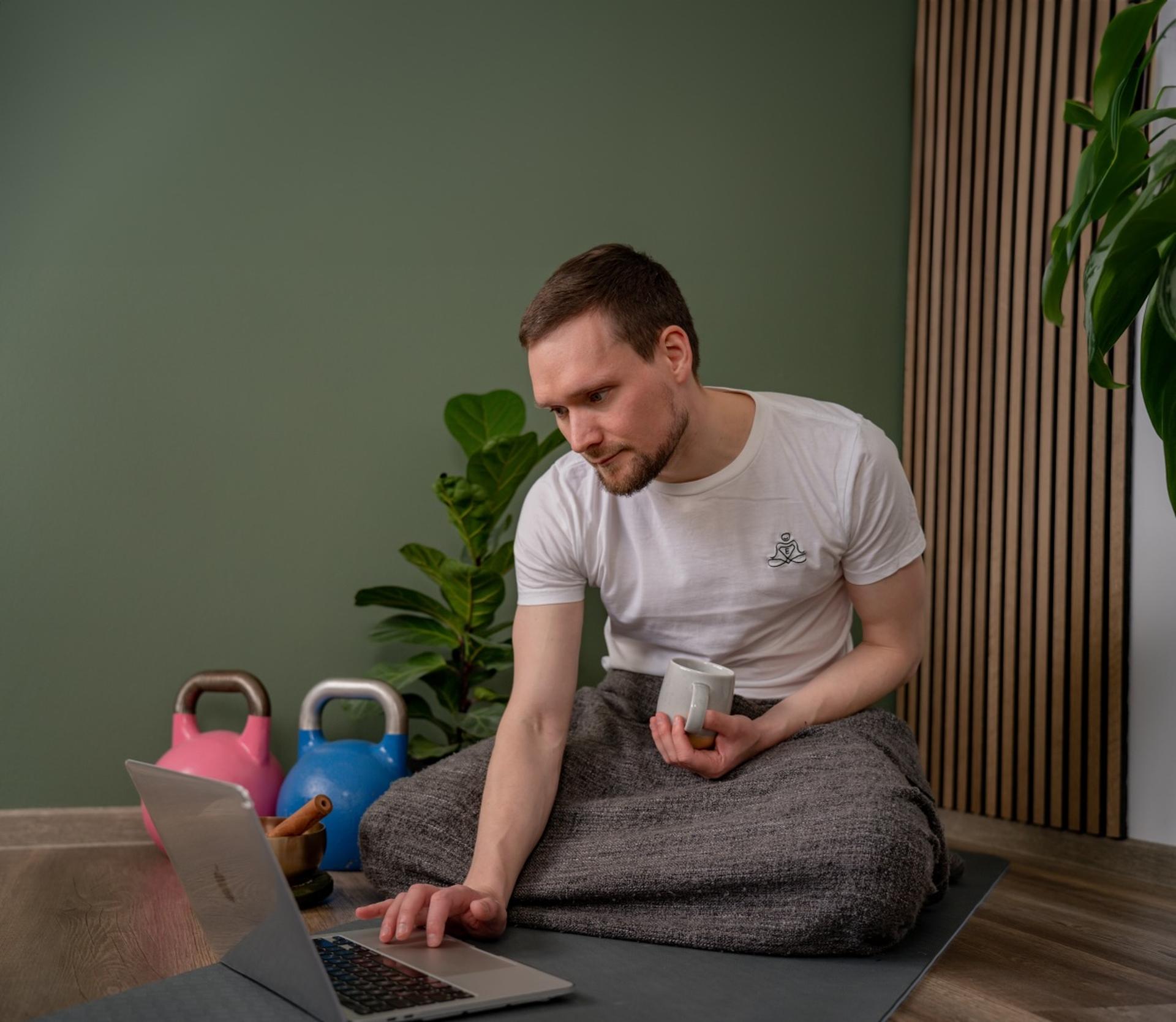Entspannungstrainer Johannes bei Online-Coaching Session mit Laptop und Kaffee, umgeben von Trainingsgeräten und Zimmerpflanzen in entspannender Atmosphäre