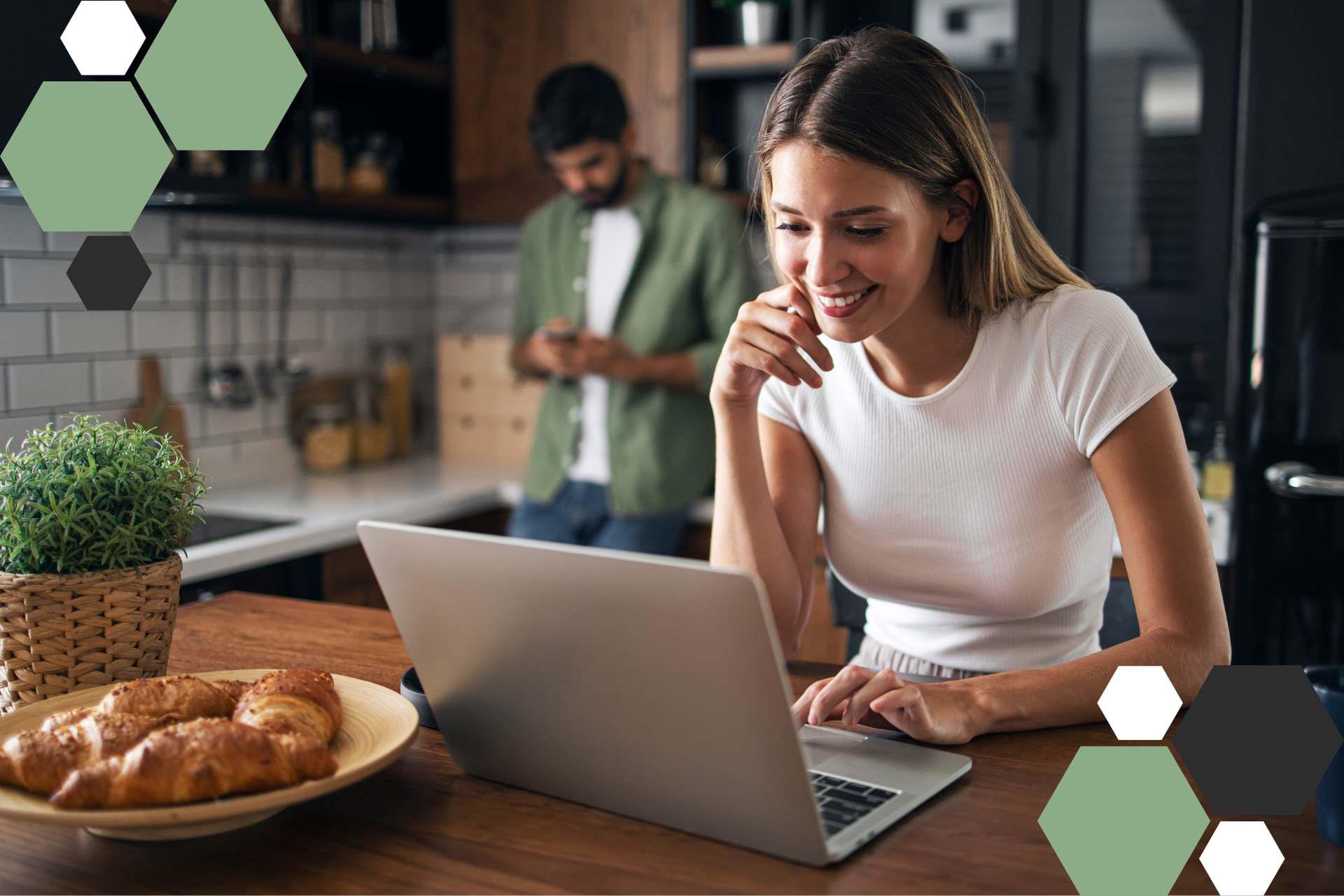 Eine Frau sitzt in der Küche mit dem Laptop. Ein Mann ist im Hintergrund und schaut auf sein Handy.