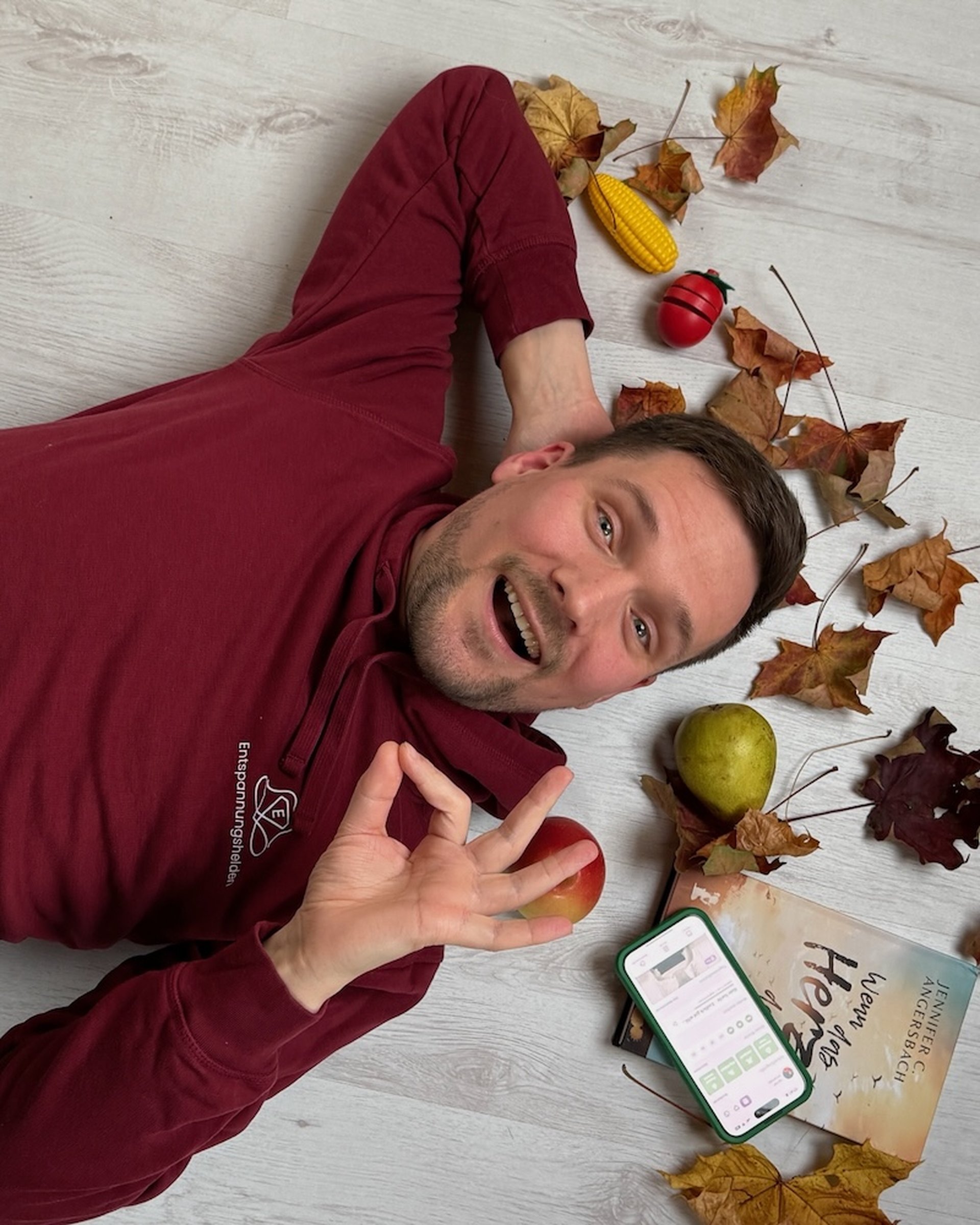 Johannes liegt in einem kastanienbraunen Kapuzenpulli mit Herbstblättern, Äpfeln und einem Telefon auf dem Boden. Er lächelt und macht mit der Hand eine „OK“-Geste.