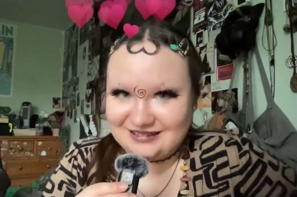Young woman with spiral drawn between eyes and pink hearts above head talking into a fluffy mic in room