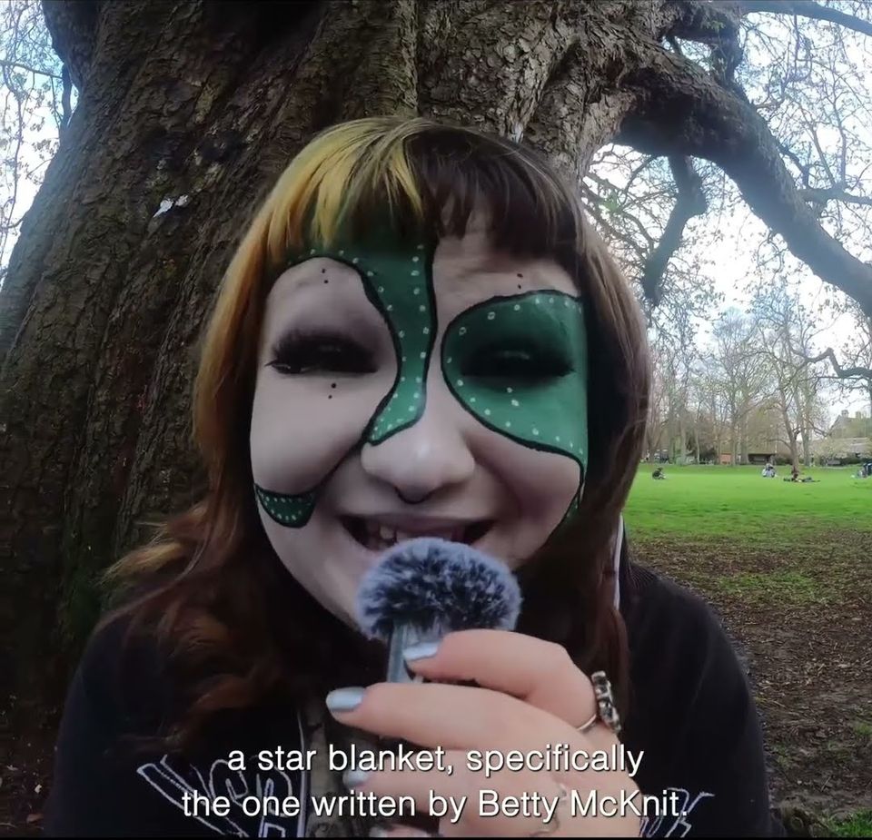 Young woman with white and green paint on face talking into a fluffy mic outside with subtitle "a star blanket, specifically the one written by Betty McKnit."