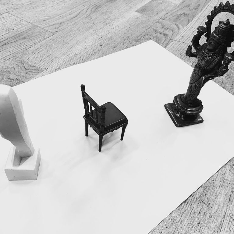 A small chair on a white sheet, flanked by a white abstract sculpture on the left and a dark statue on the right, on a wooden floor.