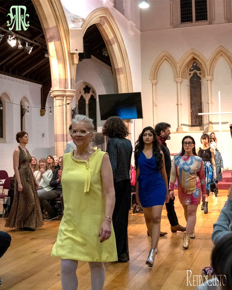 A diverse group of models walks down a church aisle runway, showcasing various colorful outfits during a fashion show.