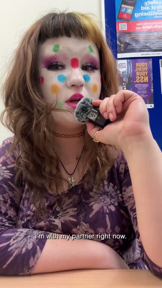 Young woman white white paint and coloured spots on face talking into a fluffy mic with audio caption "I'm with my partner right now"