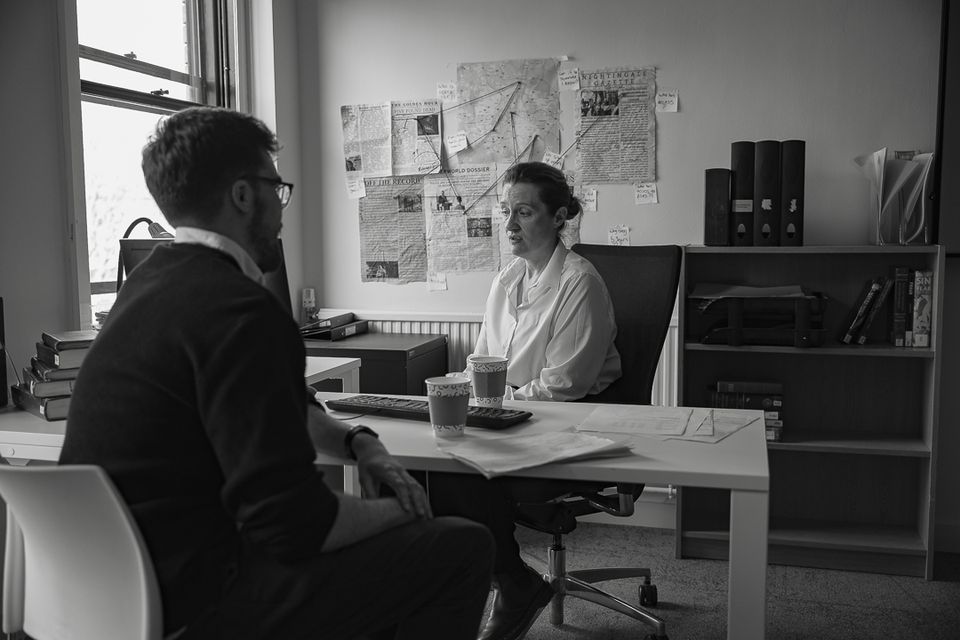 A man and woman engaged in conversation in an office. Papers, books, and pinned documents surround them. A window offers natural light.