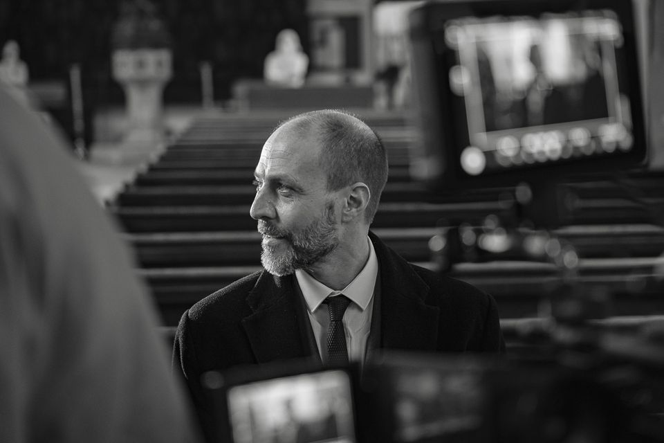 Black and white image of a man in a suit, looking to the side, seated in a church, with camera equipment visible in the foreground.