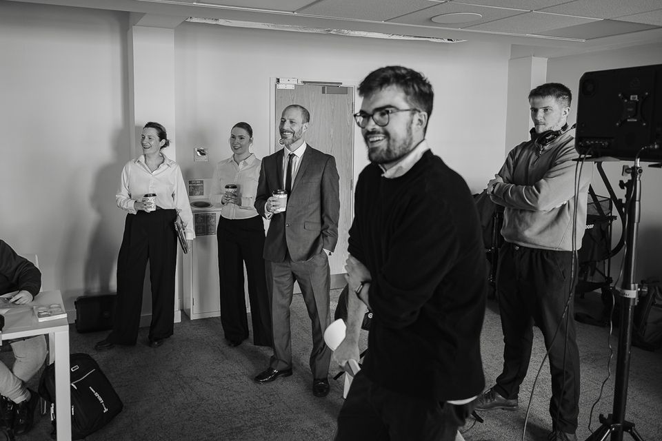 A group of five people casually chatting and smiling in a room, some holding coffee cups. A camera setup is visible on the right. Black and white.