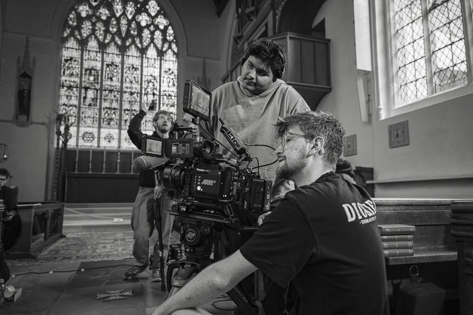 Film crew operates a camera inside a church, with a large stained glass window in the background.