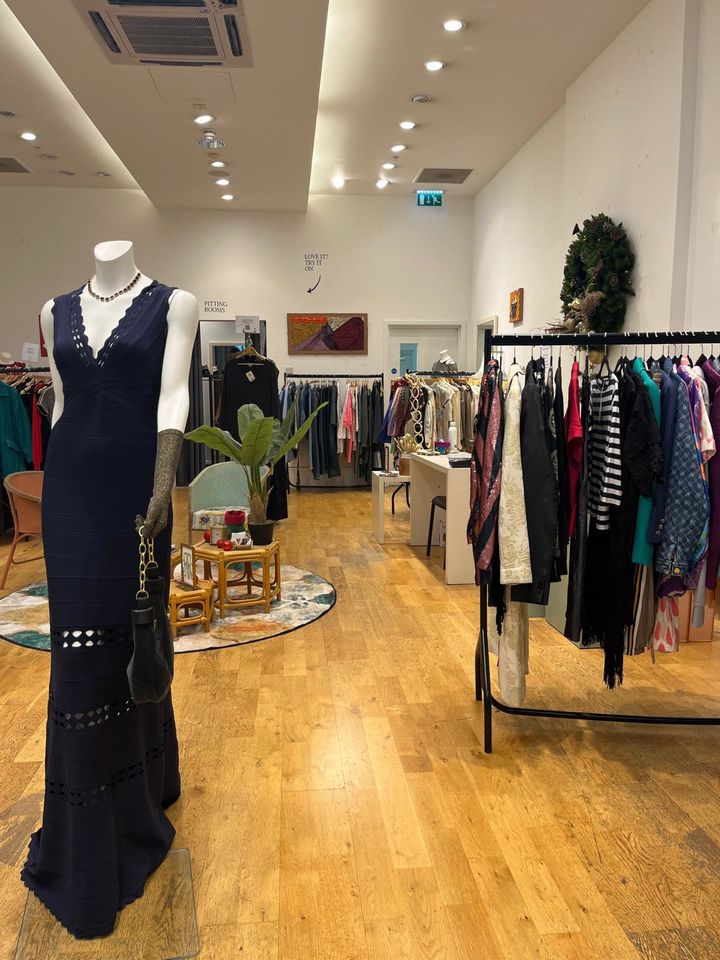 Boutique interior with a mannequin in a black dress, various colorful clothes on racks, and a small seating area with plants and decor.