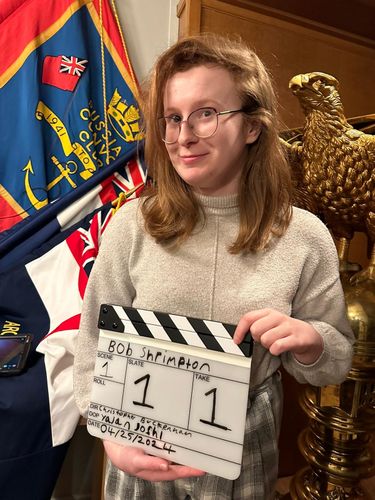 Eleanor Lynch holding a clapperboard in front of Royal Navy regalia