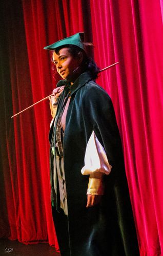 Female actor in a green robe and cap holds a long arrow, standing in front of red curtains on stage.