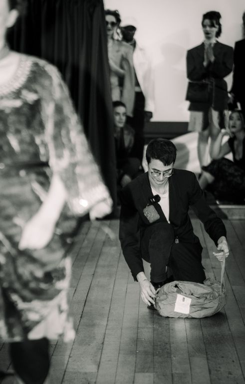 Cosmin Diaconu in a suit kneeling on a wooden floor, interacting with a bag. Blurred figure in the foreground, audience in the background. Black and white.