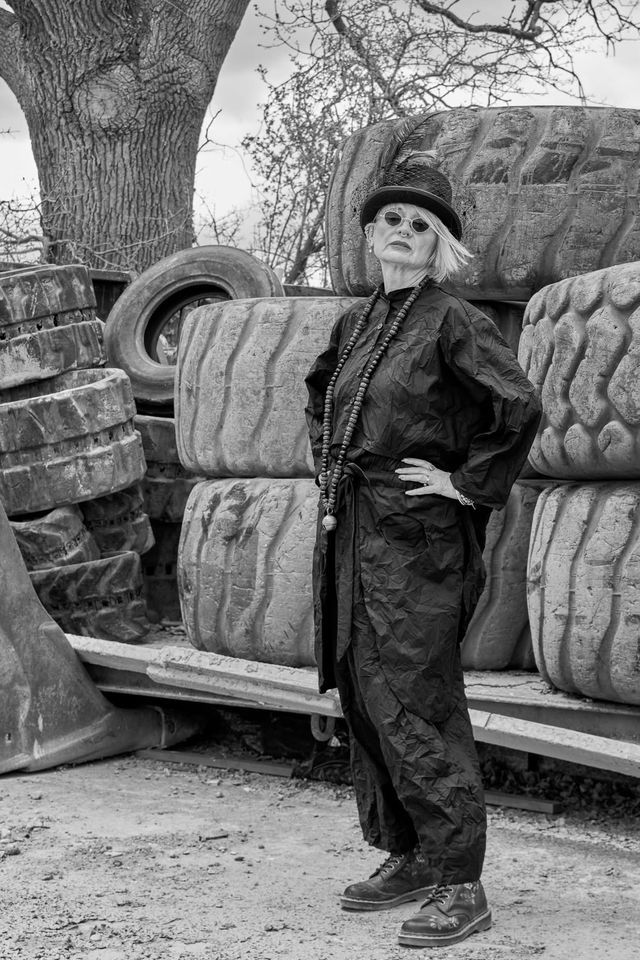 Black and white photo of female model wearing sunglasses and black boiler suit in front of pile of large tyres