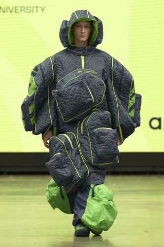 Model in oversized, quilted outfit with green accents and multiple pockets, walking on a runway with a yellow background.