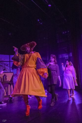 Actor in a vibrant orange costume dances energetically on stage, followed by others in more casual attire, under purple lighting.