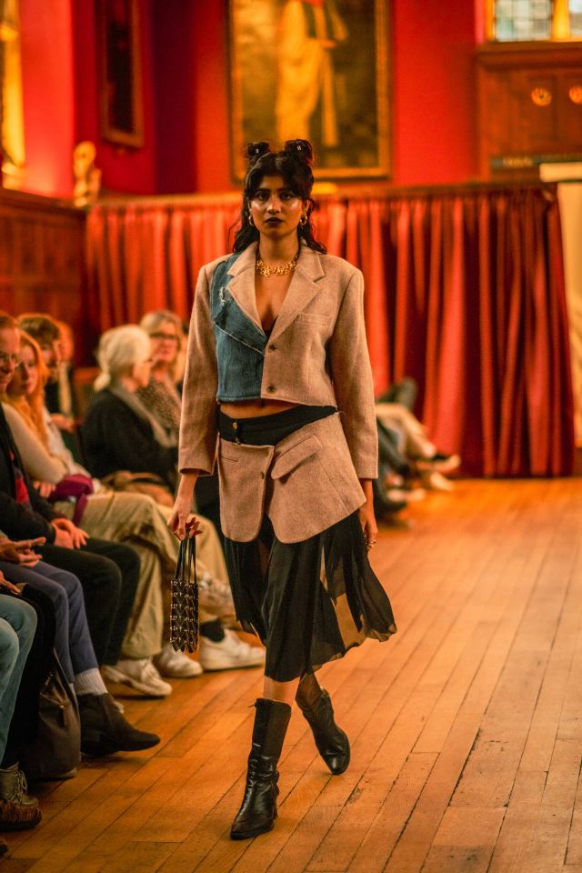 Female model walking on a wooden floor in a stylish blazer and skirt, holding a handbag. Audience seated on either side, with red curtains in the background.