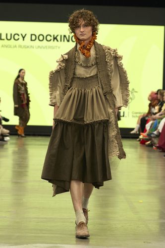 Model in layered, earth-toned outfit with ruffles and scarf walks runway; bright yellow backdrop with text and seated audience.