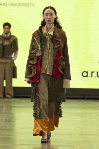 Model in a layered, earthy-toned outfit with ruffled details walks on a runway against a yellow backdrop.
