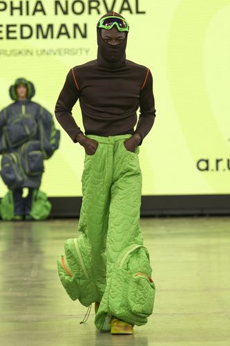 Model in a brown top and green quilted pants with oversized pockets walks on a runway. Background features a yellow screen with text.