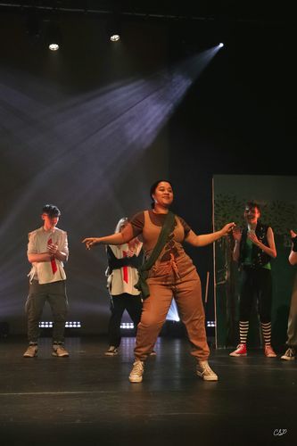 Female actor in brown jumpsuit dances energetically on stage with others in background under dramatic lighting.