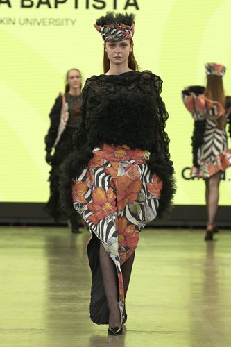 Model in a black textured top and floral-patterned skirt walks the runway against a bright yellow backdrop.