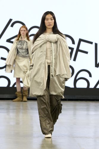 Female model wearing beige knotted coat over beige top and dark green trousers on GFW2026 catwalk