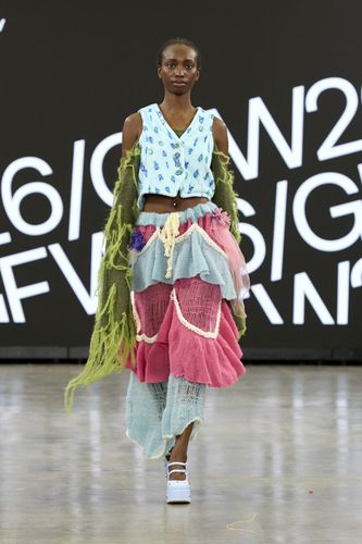 Female model wearing blue sleeveless top, blue and red sheer skirt and green vest with long flowing sides on GFW2026 catwalk