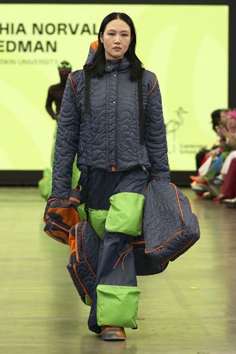 Model in a quilted blue and green outfit with orange accents walks a runway against a bright yellow backdrop.