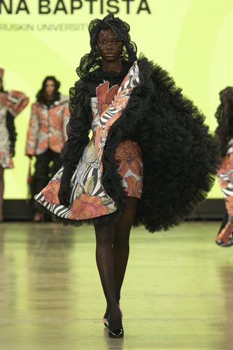 Model in a floral dress with a large black ruffled cape walks the runway against a bright yellow backdrop.
