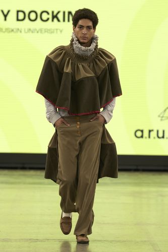 Model wearing a layered brown outfit with ruffled collar and wide pants walks on a runway. Bright yellow background with text.