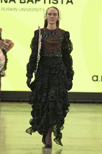 Model in a ruffled black dress with sheer sleeves and a floral design, wearing a netted headpiece, walks on a runway with a yellow background.