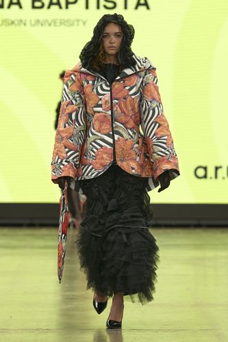 Model in a floral and zebra-print jacket over a black ruffled dress, walking on a runway with a bright yellow backdrop.