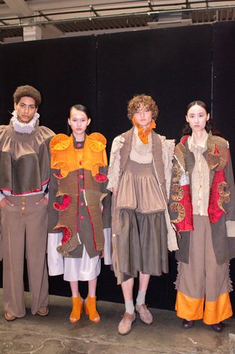 Four models in colorful, avant-garde outfits with layered textures and bold patterns pose against a black backdrop.