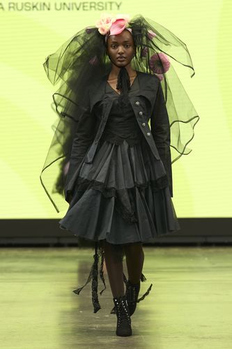 Model in a black layered dress with a large black veil adorned with pink flowers, walking on a runway.