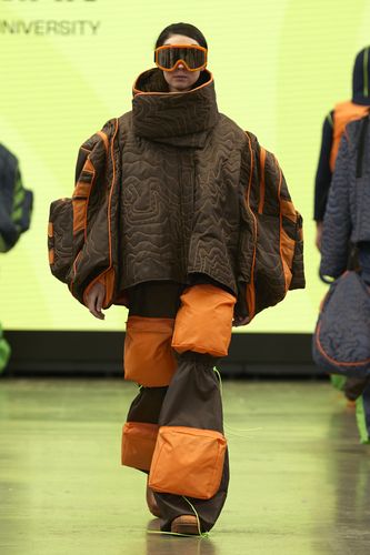 Model in a futuristic, oversized brown and orange quilted outfit with matching visor sunglasses, walking on a runway.