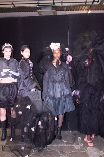 Four models in avant-garde black outfits with pink floral accents pose against a dark backdrop.