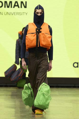 Model wearing a navy hooded outfit with an orange vest and green padded pants on a runway with a yellow background.