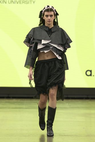 Model in layered black and pink outfit with ruffled top and skirt, wearing knee-high boots, walks on runway with yellow background.