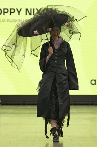 Model in a black outfit and large hat with veil, holding an umbrella adorned with pink flowers, walks on a runway with a bright yellow backdrop.