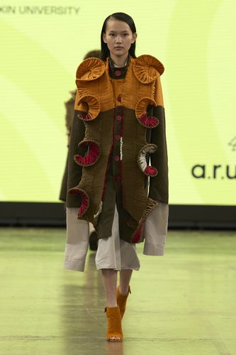 Model in a unique, textured outfit with orange and brown ruffles walks on a runway with a bright yellow background.
